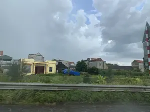 Bức ảnh chụp quang cảnh một khu dân cư ở vùng nông thôn hoặc ngoại ô, có thể được chụp từ một phương tiện đang di chuyển. Ở tiền cảnh là một lan can đường bộ bằng kim loại và thảm cỏ xanh tươi. Phía sau lan can là nhiều ngôi nhà với các kiểu dáng khác nhau, bao gồm cả nhà mới sơn màu vàng và nhà cũ hơn với tường xi măng. Một số ngôi nhà có bồn chứa nước bằng inox trên mái. Cây cối và một tấm bạt màu xanh cũng có thể được nhìn thấy giữa các ngôi nhà. Ở phía xa bên phải, có một cột báo hiệu sọc đỏ và trắng, có thể là biển báo đường sắt. Bầu trời u ám và đầy mây.。（日本語訳）。田舎または郊外の住宅地の風景を写した写真で、移動中の乗り物から撮影された可能性があります。手前には金属製のガードレールと青々とした芝生があります。ガードレールの後ろには、新しく黄色に塗られた家やセメント壁の古い家など、さまざまな様式の家が多数あります。いくつかの家は屋根にステンレス製の貯水タンクを設置しています。家々の間には木々や青い防水シートも見えます。遠くの右側には、赤と白の縞模様の標識柱があり、鉄道標識かもしれません。空はどんよりと曇っています。