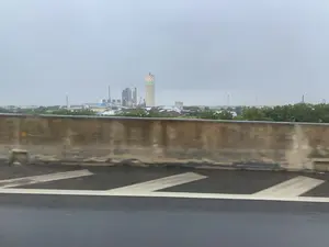 Bức ảnh được chụp từ một phương tiện đang di chuyển, có thể là trên cầu vượt hoặc đường cao tốc, nhìn ra một khu công nghiệp rộng lớn dưới bầu trời u ám. Ở phía xa, có một tổ hợp nhà máy với nhiều công trình, ống khói và silo. Nổi bật là một tháp hoặc ống khói bê tông cao, trên đỉnh có hoa văn ô cờ và dòng chữ có thể đọc được là 'B.ĐNB'. Phía trước khu nhà máy là một hàng cây xanh và có thể thấy các đống nguyên liệu (có thể là than đá) cùng một chiếc máy xúc. Phần tiền cảnh của bức ảnh là lan can bê tông của cây cầu và mặt đường nhựa có vạch kẻ trắng, bị mờ đi do tốc độ di chuyển.。（日本語訳）。この写真は、高架橋または高速道路上と思われる移動中の乗り物から撮影されたもので、曇り空の下に広がる広大な工業地帯を眺めています。遠方には、多くの建造物、煙突、サイロを備えた工場複合体があります。目立っているのは高いコンクリートの塔または煙突で、その頂上には市松模様と「B.ĐNB」と読み取れる文字があります。工場地帯の手前には緑の木々が並んでおり、原料の山（おそらく石炭）と一台の掘削機が見えます。写真の前景には、橋のコンクリート製の手すりと白い線が引かれたアスファルトの路面があり、移動速度によってぼやけています。