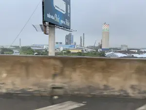 Bức ảnh chụp quang cảnh một khu công nghiệp lớn, có thể là một nhà máy xi măng, nhìn từ một con đường trên cao hoặc cầu vượt. Ở tiền cảnh, một bức tường chắn bê tông bị mờ do chuyển động. Ở phía sau là một khu phức hợp nhà máy rộng lớn dưới bầu trời u ám, bao gồm các tòa nhà sản xuất, ống khói, và các tháp cao. Một tháp xi-lô bê tông nổi bật với họa tiết bàn cờ ở trên cùng và có dòng chữ 'NB.DNB'. Có thể nhìn thấy các cần cẩu, hệ thống băng tải, và những đống nguyên vật liệu lớn, một số được che phủ bởi bạt trắng. Một biển quảng cáo lớn ở bên trái có dòng chữ 'ĐIỀU HÒA Chất' và 'DỊCH VỤ Nhất', quảng cáo cho một sản phẩm điều hòa không khí.。（日本語訳）。写真は、高架道路または陸橋から見た、大きな工業団地、おそらくセメント工場の光景を撮影したものです。前景には、動きによってぼやけたコンクリートの防護壁があります。後方には、曇り空の下に広大な工場複合施設があり、生産建屋、煙突、そして高い塔が含まれています。一つのコンクリートのサイロタワーが目立っており、その上部にはチェッカー盤模様があり、「NB.DNB」という文字が書かれています。クレーン、コンベアシステム、そして大きな原材料の山が見え、その一部は白いシートで覆われています。左側にある大きな広告看板には「ĐIỀU HÒA Chất」と「DỊCH VỤ Nhất」という文字があり、エアコン製品を宣伝しています。
