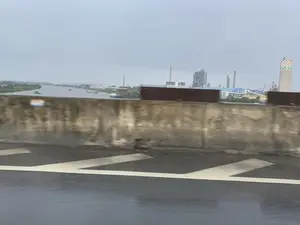 Bức ảnh được chụp từ một phương tiện đang di chuyển trên cầu hoặc đường trên cao, nhìn ra một khu công nghiệp nằm bên cạnh một con sông rộng. Bầu trời u ám và mặt đường có vẻ ẩm ướt. Dưới sông, có một vài chiếc tàu và sà lan đang hoạt động, có khả năng là để vận chuyển hàng hóa. Ở phía xa, khu công nghiệp có nhiều nhà máy, ống khói, và một tháp chứa hình trụ nổi bật. Trên tháp này có dòng chữ "NB.DN" và phần đỉnh được trang trí bằng hoa văn ca rô đỏ và trắng. Ở tiền cảnh, rào chắn bê tông của cây cầu và vạch kẻ đường bị mờ do chuyển động.。（日本語訳）。この写真は、橋または高架道路を走行中の乗り物から撮影されたもので、広い川の隣にある工業団地を眺めている。川には、数隻の船やはしけが航行しており、おそらく貨物を輸送するためだろう。遠くには、工業団地に多くの工場、煙突、そして目立つ円筒形の貯蔵塔がある。この塔には「NB.DN」という文字があり、上部は赤と白の市松模様で装飾されている。手前には、橋のコンクリートの防護柵と車線が、動きによってぼやけている。