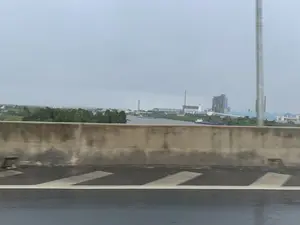 Bức ảnh được chụp từ trên một cây cầu hoặc đường cao tốc, nhìn qua dải phân cách bê tông. Mặt đường có vẻ ướt, cho thấy trời có thể vừa mưa. Ở phía xa là một con sông rộng, trên đó có một vài chiếc tàu và sà lan đang di chuyển, có khả năng là để vận chuyển hàng hóa. Bên kia bờ sông là một khu công nghiệp quy mô lớn với nhiều nhà máy, nhà xưởng có mái vòm màu xanh, và các ống khói cao. Một số tòa nhà trong khu công nghiệp trông giống như đang trong quá trình xây dựng. Bầu trời u ám và có nhiều mây.。（日本語訳）。写真は橋または高速道路の上から、コンクリート製の中央分離帯越しに撮影されたものです。路面は濡れているように見え、雨が降ったばかりの可能性があります。遠くには広い川があり、その上を数隻の船やはしけが移動しており、貨物を輸送している可能性があります。川の対岸には大規模な工業地帯があり、多くの工場、青いドーム型の屋根を持つ作業場、そして高い煙突があります。工業地帯の一部の建物は建設中のように見えます。空はどんよりとしており、雲が多いです。