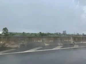 Bức ảnh được chụp từ một phương tiện đang di chuyển, có thể là trên đường cao tốc hoặc cầu cạn, vào một ngày trời u ám. Ở phía trước là dải phân cách bằng bê tông và mặt đường nhựa ướt, có các vạch kẻ đường màu trắng. Nhìn qua dải phân cách, phía xa là một khu vực cảnh quan kết hợp giữa nông thôn và công nghiệp. Có thể thấy cây cối xanh tươi, xen kẽ là một vài ngôi nhà và các công trình nhà máy, nhà xưởng với những ống khói cao vươn lên bầu trời xám xịt. Bầu trời đầy mây, cho thấy thời tiết không thuận lợi, có thể đang hoặc vừa mới mưa.。（日本語訳）。写真は、移動中の乗り物から、おそらく高速道路か高架橋の上で、曇りの日に撮影されたものです。手前にはコンクリートの中央分離帯と、白い車線のある濡れたアスファルトの路面があります。中央分離帯の向こう、遠方には田園と工業が混在した景観が広がっています。緑の木々が見え、その中にいくつかの家や、灰色の空に向かって伸びる高い煙突を持つ工場や作業場が点在しています。空は雲でいっぱいで、天気が良くないことを示しており、雨が降っているか、あるいは降った直後かもしれません。