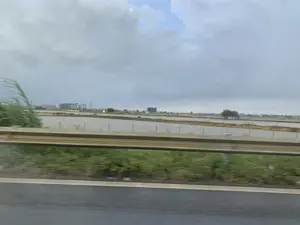 Bức ảnh được chụp từ một phương tiện đang di chuyển, nhìn ra khung cảnh vùng nông thôn ven đường. Ở phía trước, một dải lan can đường bằng kim loại chạy ngang qua khung hình. Phía sau lan can là một vùng nông địa rộng lớn, bao gồm nhiều cánh đồng hoặc đầm ngập nước, được phân chia bởi các bờ đất. Cảnh quan này có thể là ruộng lúa đang trong giai đoạn chuẩn bị gieo cấy hoặc các đầm nuôi trồng thủy sản. Rải rác trong khu vực là cây cối, một vài ngôi nhà, các cột điện cao thế và một công trình nhỏ trông giống miếu thờ. Bầu trời u ám, đầy mây xám.。（日本語訳）。写真は移動中の乗り物から撮影されたもので、道端の田園風景を眺めている。手前には、金属製のガードレールがフレームを横切っている。ガードレールの後ろには広大な農地が広がっており、土手で区切られた多くの田んぼや水で満たされた沼地が含まれている。この風景は、田植えの準備段階にある水田か、あるいは水産養殖の池である可能性がある。空はどんよりとしており、灰色の雲で覆われている。