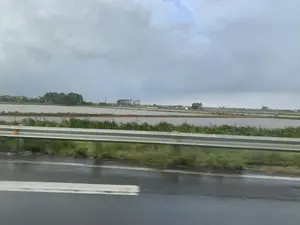 Bức ảnh được chụp từ một phương tiện đang di chuyển, nhìn ra khung cảnh nông thôn bên đường. Ở phía trước là một con đường nhựa ướt và một dải lan can bằng kim loại, cho thấy có thể trời vừa mưa. Phía sau lan can là một cánh đồng rộng lớn ngập nước, có khả năng là ruộng lúa đang trong giai đoạn chuẩn bị gieo cấy. Xa xa phía chân trời là những lùm cây, một vài ngôi nhà và các cột điện. Bầu trời u ám, bao phủ bởi những đám mây xám. Không có chữ viết hay biển hiệu nào có thể đọc được trong ảnh.。（日本語訳）。手前には濡れたアスファルトの道路と金属製のガードレールがあり、雨が降ったばかりの可能性があることを示しています。ガードレールの後ろには水で満たされた広大な畑があり、田植えの準備段階にある水田である可能性があります。遠くの地平線には、木々の茂み、いくつかの家、そして電柱があります。空はどんよりとしており、灰色の雲に覆われています。写真には読み取れる文字や看板はありません。