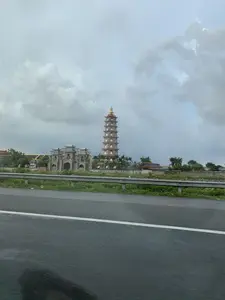 Bức ảnh được chụp từ một chiếc xe đang di chuyển trên đường, nhìn ra một công trình kiến trúc tôn giáo lớn. Ở phía trước là mặt đường nhựa ướt và một hàng rào chắn kim loại. Phía sau hàng rào là một quần thể chùa Phật giáo. Công trình nổi bật nhất là một ngọn bảo tháp cao, có nhiều tầng, hình bát giác, với các hốc tường đặt tượng Phật ở mỗi tầng. Bên cạnh ngọn tháp là một công trình cổng tam quan lớn dường như đang trong quá trình xây dựng. Xung quanh có cây cối, bao gồm cả cây cọ, và một số lá cờ Phật giáo màu vàng đang được treo. Bầu trời u ám và đầy mây xám.。（日本語訳）。手前には濡れたアスファルトの路面と金属製のガードレールがある。ガードレールの後ろには仏教寺院の複合施設がある。最も目立つ建物は、高く、多層で、八角形の仏塔であり、各層の壁龕には仏像が安置されている。塔の隣には、建設中と思われる大きな三門がある。空は薄暗く、灰色の雲で覆われている。