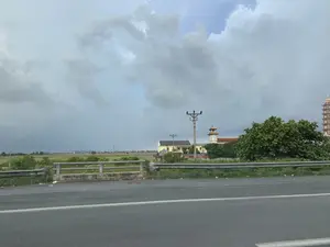 Bức ảnh chụp phong cảnh vùng nông thôn nhìn từ một con đường có lan can bảo vệ. Phía xa là một cánh đồng lúa rộng lớn. Nổi bật trong khung cảnh là một quần thể kiến trúc dường như là một ngôi chùa, với một tòa tháp đồng hồ và một bảo tháp nhiều tầng có mái cong đặc trưng. Bên cạnh đó là một ngôi nhà dân và nhiều cây xanh. Có các cột điện chạy dọc theo khu vực. Bầu trời u ám và đầy mây. Không có văn bản hay chữ viết nào có thể nhận dạng được trong ảnh.。（日本語訳）。ガードレールのある道から見た、田舎の風景を撮影した写真です。遠くには広大な水田が広がっています。風景の中で際立っているのは、寺院と思われる建築群で、時計塔と、特徴的な反りのある屋根を持つ多層の仏塔があります。その隣には民家一軒と多くの緑の木々があります。その地域に沿って電柱が立っています。空はどんよりとしており、雲に覆われています。写真の中には、認識できるテキストや文字はありません。