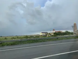 Bức ảnh chụp phong cảnh nông thôn từ một phương tiện đang di chuyển trên đường. Ở tiền cảnh là một phần của con đường nhựa và hàng rào chắn ven đường. Phía sau hàng rào là một quần thể kiến trúc tôn giáo, dường như là một ngôi chùa, với các tòa nhà có mái ngói cong truyền thống. Nổi bật trong quần thể là một bảo tháp cao nhiều tầng và một tháp khác có gắn đồng hồ. Xung quanh chùa là cây cối xanh tươi. Xa hơn nữa là những cánh đồng lúa rộng lớn. Bầu trời u ám và bao phủ bởi những đám mây xám dày đặc, báo hiệu thời tiết xấu.。（日本語訳）。道路を移動中の乗り物から田園風景を撮影した写真。手前には、アスファルト道路の一部と道路脇のガードレールが写っている。ガードレールの後ろには、伝統的な反りのある瓦屋根を持つ建物がある、寺院と思われる宗教的な建築物群がある。その建築物群の中で目立っているのは、高い多層の仏塔と、時計が取り付けられたもう一つの塔である。寺院の周りには青々とした木々がある。さらにその奥には広大な水田が広がっている。空はどんよりとしており、厚い灰色の雲に覆われ、悪天候を予感させる。