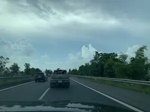 Hình ảnh được chụp từ bên trong một chiếc ô tô đang di chuyển trên một con đường cao tốc có nhiều làn xe. Phía trước có một chiếc xe bán tải màu be hiệu Ford Ranger, trên thùng xe đang chở ít nhất một chiếc xe máy. Bên cạnh xe bán tải là một chiếc ô tô SUV màu đen. Hai bên đường có cây cối xanh tươi và lan can bảo vệ. Bầu trời u ám và có nhiều mây.。（日本語訳）。複数車線のある高速道路を走行中の車内から撮影された画像です。前方にはフォード・レンジャーのベージュ色のピックアップトラックがおり、その荷台には少なくとも一台のバイクを積んでいます。ピックアップトラックの隣には、黒色のSUV車がいます。道路の両側には、青々とした木々とガードレールがあります。空はどんよりとしており、雲が多いです。