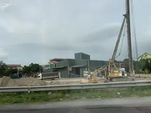Cảnh quan một công trường xây dựng ven đường. Ở trung tâm là các máy móc hạng nặng, bao gồm một máy xúc màu vàng và một giàn máy đóng cọc cao. Trên thân máy đóng cọc có thể đọc được chữ 'K2' và có vẻ như là 'PHUC VY GROUP'. Nền đất của công trường là đất và cát, có nhiều đống đất được vun lên. Phía sau khu vực thi công là một tòa nhà lớn giống như nhà xưởng hoặc kho bãi với tường và mái tôn màu xanh. Một chiếc xe tải màu trắng có bạt che đang đỗ gần đó. Xa hơn trong hậu cảnh là các ngôi nhà dân, cây cối và một ngôi nhà sơn màu xanh lá cây. Bầu trời u ám và đầy mây. Phía trước có một lan can bảo vệ bằng kim loại, cho thấy ảnh được chụp từ trên một con đường.。（日本語訳）。道路沿いの建設現場の風景。中央には、黄色いショベルカーと高い杭打ち機を含む重機があります。杭打ち機の本体には「K2」という文字と、「PHUC VY GROUP」と思われる文字が読み取れます。建設現場の地面は土と砂で、多くの土の山が積み上げられています。工事エリアの後ろには、青いトタンの壁と屋根を持つ、工場か倉庫のような大きな建物があります。近くには幌で覆われた白いトラックが駐車しています。さらに奥の背景には、民家、木々、そして緑色に塗られた家があります。空はどんよりとしており、雲に覆われています。手前には金属製のガードレールがあり、写真が道路上から撮影されたことを示しています。