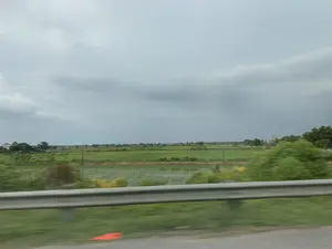 Bức ảnh được chụp từ một phương tiện đang di chuyển, nhìn ra khung cảnh vùng nông thôn. Ở phía trước là một lan can bảo vệ bằng kim loại ven đường, cho thấy ảnh được chụp từ một con đường lớn hoặc cao tốc. Phía sau lan can là một cánh đồng lúa xanh bạt ngàn, xen kẽ là những ao nước. Xa xa có thể thấy một vài ngôi nhà của người dân và những hàng cây. Có các cột điện chạy dọc theo cánh đồng. Bầu trời u ám, đầy mây xám, báo hiệu thời tiết sắp có mưa.。（日本語訳）。移動中の乗り物から撮影された写真で、田園風景が写っている。手前には道路脇の金属製のガードレールがあり、写真が主要な道路または高速道路から撮影されたことを示している。ガードレールの後ろには、広大な緑の水田が広がり、池が点在している。田んぼに沿って電柱が立っている。空はどんよりとして灰色の雲に覆われ、もうすぐ雨が降る気配がする。