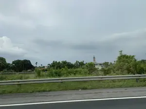 Bức ảnh ghi lại khung cảnh vùng nông thôn Việt Nam, được chụp từ một phương tiện đang di chuyển trên đường lớn, có thể là đường cao tốc. Ở tiền cảnh là dải hộ lan bằng kim loại và một phần mặt đường, hình ảnh bị mờ do chuyển động. Phía sau dải hộ lan là một khu vực có nhiều cây cối xanh tươi. Ở phía xa, nổi bật lên trên nền cây xanh là tháp chuông của một nhà thờ. Bầu trời u ám và đầy mây. Không có hoạt động cụ thể nào của con người được quan sát rõ. Không có văn bản hay chữ viết nào có thể đọc được trong ảnh.。（日本語訳）。この写真はベトナムの田園風景を記録したもので、大きな道路、おそらく高速道路を移動中の乗り物から撮影されたものです。前景には金属製のガードレールと路面の一部があり、動きによって画像がぼやけています。ガードレールの後ろには、緑豊かな木々がたくさんあるエリアが広がっています。遠くには、緑の木々を背景にして教会の鐘楼が際立っています。空はどんよりとしており、雲に覆われています。特定の人間の活動ははっきりと観察されません。写真の中には読み取れるテキストや文字はありません。