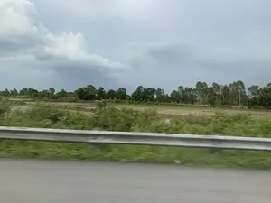 Bức ảnh được chụp từ một phương tiện đang di chuyển, nhìn ra khung cảnh nông thôn ven đường. Ở tiền cảnh, có một lan can đường bằng kim loại và lề đường cỏ xanh bị mờ do chuyển động. Phía sau lan can là một cánh đồng rộng lớn, có thể là ruộng lúa. Một số khu vực của cánh đồng đã được cày xới hoặc thu hoạch, để lộ đất nâu và một ít nước đọng. Xa hơn là một hàng cây xanh rậm rạp. Bầu trời u ám và đầy mây, báo hiệu một ngày không có nắng. Không có bất kỳ văn bản, biển báo hay hoạt động cụ thể nào của con người được ghi lại.。（日本語訳）。写真は移動中の乗り物から撮影されたもので、道端の田園風景を望んでいます。前景には、金属製のガードレールと、動きによってぼやけた緑の草の路肩があります。ガードレールの後ろには広大な畑が広がっており、おそらく水田でしょう。畑の一部は耕されたり収穫されたりしており、茶色い土と少しの水たまりが見えます。その向こうには、緑豊かな木々が立ち並んでいます。空はどんよりと曇っており、晴れていない日であることを示しています。文字、標識、または特定の人間活動は記録されていません。