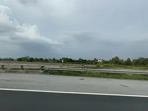 Bức ảnh chụp phong cảnh nông thôn ven đường. Ở tiền cảnh là một phần của con đường nhựa và lan can bảo vệ. Phía sau lan can là những cánh đồng lúa nước rộng lớn, mặt ruộng ngập nước, có thể đang trong giai đoạn chuẩn bị cho vụ mùa mới. Xa xa là những hàng cây xanh và một vài ngôi nhà của người dân, nổi bật có một ngôi nhà mái ngói đỏ. Bầu trời u ám, nhiều mây, báo hiệu thời tiết không nắng. Không có chữ viết nào có thể nhận dạng được.。（日本語訳）。道端の田舎の風景を撮った写真です。手前には、アスファルトの道路の一部とガードレールがあります。ガードレールの後ろには広大な水田が広がっており、田んぼの表面は水で満たされ、おそらく新しい作付けシーズンの準備段階にあるようです。遠くには緑の木々の列といくつかの民家があり、中でも赤い瓦屋根の家が目立っています。空はどんよりとして雲が多く、晴れていない天気であることを示しています。認識できる文字はありません。