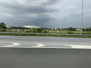 Bức ảnh ghi lại quang cảnh nhìn từ một phương tiện đang di chuyển trên đường cao tốc. Ở tiền cảnh là mặt đường nhựa với vạch kẻ đường màu trắng và một dải hộ lan kim loại. Bên kia đường là một cánh đồng, trên đó có một vài ngôi mộ. Ở phía xa là cảnh quan nông thôn với cây cối, nhà dân và một tòa nhà lớn, hiện đại màu trắng. Bầu trời u ám, bao phủ bởi những đám mây xám dày đặc, báo hiệu thời tiết xấu hoặc sắp có mưa.。（日本語訳）。写真は、高速道路を走行中の乗り物から見た光景を捉えている。前景には、白い車線と金属製のガードレールがあるアスファルトの路面がある。道路の向こう側には野原があり、そこにはいくつか墓がある。遠くには、木々、民家、そして大きな白い近代的な建物がある田園風景が広がっている。空はどんよりとしており、厚い灰色の雲に覆われ、悪天候または雨が降りそうなことを示している。