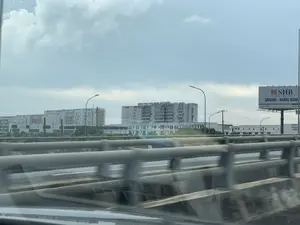 Bức ảnh được chụp từ một phương tiện đang di chuyển trên đường cao tốc hoặc cầu vượt, nhìn ra ngoài qua lan can. Do xe đang chạy nên hình ảnh lan can ở tiền cảnh bị mờ. Ở phía xa là một khu vực có nhiều tòa nhà lớn, hiện đại. Nổi bật là một tòa nhà lớn có dòng chữ "BỆNH VIỆN HỮU NGHỊ VIỆT ĐỨC" trên nóc. Bên phải có một biển quảng cáo lớn của ngân hàng SHB với nội dung: "SHB", "Solid partners, flexible solutions", "SAIGON - HANOI BANK", "Hotline: *6688", và một phần địa chỉ website. Khung cảnh chung cho thấy một khu vực đang phát triển với các công trình lớn, có cột đèn đường và bầu trời nhiều mây.。（日本語訳）。この写真は、高速道路または高架橋を走行中の乗り物から、欄干越しに外を向いて撮影されたものです。車が走行中であるため、前景の欄干の画像はぼやけています。遠方には、多くの大きくて近代的な建物があるエリアが広がっています。屋上に「BỆNH VIỆN HỮU NGHỊ VIỆT ĐỨC」という文字がある大きな建物が目立っています。右側にはSHB銀行の大きな広告看板があり、その内容は「SHB」、「Solid partners, flexible solutions」、「SAIGON - HANOI BANK」、「ホットライン：*6688」、そしてウェブサイトアドレスの一部です。全体的な風景は、大きな建築物、街灯の柱、そして曇り空がある、発展中の地域を示しています。