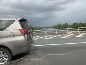 Bức ảnh được chụp từ bên trong một chiếc ô tô đang di chuyển trên cầu. Một chiếc xe ô tô khác, hiệu Toyota Innova màu bạc, đang chạy song song. Có thể nhìn thấy chữ 'INNOVA' ở phía sau xe. Khung cảnh bên ngoài là một cây cầu bắc qua một con sông rộng. Ở phía xa trên mặt sông có các cấu trúc có thể là lồng bè nuôi trồng thủy sản. Bờ sông bên kia có nhiều cây xanh. Bầu trời u ám, đầy mây xám, cho thấy thời tiết có thể sắp mưa.。（日本語訳）。写真は、橋の上を走行中の車内から撮影されたものです。別の車、銀色のトヨタ・イノーバが並走しています。車の後部に「INNOVA」の文字が見えます。外の景色は、広い川にかかる橋です。遠くの川面には、水産養殖用のいかだと思われる構造物があります。対岸には緑の木々が多くあります。空はどんよりとして灰色の雲に覆われており、もうすぐ雨が降る可能性を示しています。