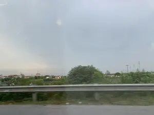 Bức ảnh được chụp từ một phương tiện đang di chuyển, cho thấy cảnh quan vùng nông thôn. Ở phía trước là một lan can đường bằng kim loại. Phía sau lan can là cây cối và thảm thực vật xanh tươi. Ở phía xa là một khu dân cư hoặc một ngôi làng với nhiều ngôi nhà mái ngói. Có thể nhìn thấy các cột điện và đường dây điện. Bầu trời u ám và có nhiều mây. Hình ảnh bị mờ do chuyển động.。（日本語訳）。この写真は移動中の乗り物から撮影されたもので、田園風景が写っている。手前には金属製のガードレールがある。ガードレールの後ろには、木々や青々とした草木がある。遠くには、多くの瓦屋根の家がある住宅地または村がある。空はどんよりとしており、雲が多い。画像は動きによってぼやけている。