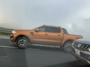 Bức ảnh chụp cảnh các phương tiện đang di chuyển trên một con đường nhựa, có thể là đường cao tốc. Đối tượng chính là một chiếc xe bán tải màu cam, có vẻ là dòng xe Ford Ranger, đang chạy song song với phương tiện chụp ảnh. Ở góc dưới bên phải, có thể thấy một phần đầu của một chiếc ô tô khác màu sẫm. Dọc bên đường có lan can an toàn, và phía xa là khung cảnh nông thôn với đồng ruộng và cây cối. Bầu trời u ám và đầy mây. Không có biển báo hay chữ viết nào có thể đọc được rõ ràng.。（日本語訳）。写真は、車両がアスファルトの道路、おそらくは高速道路を走行している光景を写したものです。主な被写体はオレンジ色のピックアップトラックで、フォード・レンジャーのモデルのようであり、撮影車両と並走しています。右下隅には、別の暗い色の車の先端部分が見えます。道路沿いには安全ガードレールがあり、遠くには田畑や木々のある田園風景が広がっています。空はどんよりとしており、雲に覆われています。はっきりと読み取れる標識や文字はありません。