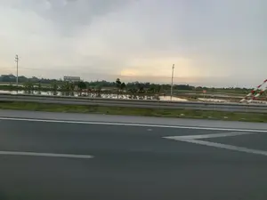 Bức ảnh chụp khung cảnh vùng nông thôn nhìn từ một con đường nhựa, có thể là đường cao tốc. Ở phía xa là những cánh đồng lúa ngập nước, phản chiếu bầu trời u ám. Dọc theo cánh đồng có cây cối, cột điện và dây điện. Có một tòa nhà ở phía xa với tấm biển quảng cáo lớn ghi chữ 'ĐẠI VIỆT NỘI THẤT TÂN CỔ ĐIỂN'. Ngoài ra còn có một biển báo nhỏ màu cam trên cánh đồng nhưng không thể đọc được nội dung. Bên phải có một vật thể sọc đỏ trắng, có thể là cọc tiêu hoặc rào chắn.。（日本語訳）。アスファルトの道路、おそらく高速道路から見た田園風景を写した写真です。遠くには水が張られた田んぼがあり、曇り空を映しています。田んぼに沿って木々、電柱、電線があります。遠くには「ĐẠI VIỆT NỘI THẤT TÂN CỔ ĐIỂN」と書かれた大きな看板のある建物があります。また、田んぼには小さなオレンジ色の看板がありますが、内容は読み取れません。右側には赤と白の縞模様の物体があり、標識杭かガードレールかもしれません。
