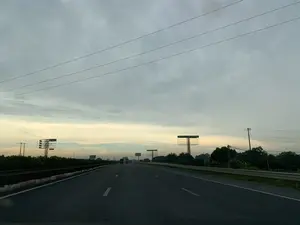 Trên một biển quảng cáo có thể đọc được dòng chữ "CHO THUÊ BẢNG QC 090 464 6699". Khung cảnh là một con đường cao tốc rộng lớn với nhiều làn xe, được chụp từ bên trong một chiếc xe đang di chuyển. Thời điểm chụp có lẽ là lúc bình minh hoặc hoàng hôn, với bầu trời u ám và có mây. Dọc hai bên đường cao tốc là dải cây xanh, lan can phòng hộ bằng kim loại và một số cột điện. Có nhiều khung biển quảng cáo lớn bằng kim loại được dựng lên, hầu hết đều đang để trống. Phía xa có thể thấy một vài phương tiện khác như ô tô và xe tải đang lưu thông trên đường.。（日本語訳）。ある広告看板に「広告看板貸出 090 464 6699」という文字が読み取れる。場面は、移動中の車内から撮影された、複数の車線がある広大な高速道路である。撮影された時間帯は、おそらく夜明けか夕暮れ時で、空は薄暗く曇っている。高速道路の両脇には、緑の木々の帯、金属製のガードレール、そしていくつかの電柱がある。