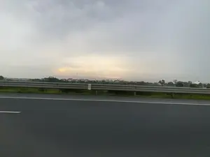 Bức ảnh ghi lại khung cảnh nhìn từ một phương tiện đang di chuyển trên một con đường lớn, có thể là đường cao tốc. Ở tiền cảnh là mặt đường nhựa màu xám với vạch kẻ đường màu trắng. Ngăn cách giữa đường và cảnh vật bên ngoài là một dải hộ lan tôn sóng bằng kim loại. Bên kia dải hộ lan là một thảm cỏ xanh. Ở phía xa, có thể thấy một khu dân cư hoặc một ngôi làng với nhiều ngôi nhà và cây cối. Bầu trời u ám, nhiều mây, có một vệt sáng ở phía chân trời. Không có văn bản nào có thể đọc được trong ảnh.。（日本語訳）。この写真は、大きな道路、おそらく高速道路を移動中の乗り物から見た風景を記録したものです。前景には、白い車線が引かれた灰色の舗装路面があります。道路と外の景色を隔てているのは、金属製の波形ガードレールです。ガードレールの向こう側には、緑の芝生があります。遠くには、多くの家や木々がある住宅地または村が見えます。空はどんよりとして雲が多く、地平線に一筋の光があります。写真の中には読み取れるテキストはありません。