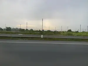 Bức ảnh được chụp từ một phương tiện đang di chuyển trên đường, nhìn ra cảnh quan ven đường. Ở phía trước là mặt đường nhựa và một dải hộ lan tôn sóng. Phía sau dải hộ lan là một khu đất rộng có cây cối và thảm thực vật. Nổi bật trên khu đất này là rất nhiều cột cao bằng kim loại, được nối với nhau bằng một hệ thống dây cáp chằng chịt, trông giống như một dàn ăng-ten vô tuyến hoặc một công trình kỹ thuật nào đó. Bầu trời u ám và đầy mây. Không có hoạt động của con người nào được nhìn thấy rõ. Không có văn bản hay chữ viết nào có thể đọc được trong ảnh.。（日本語訳）。写真は道路を移動中の乗り物から撮影されたもので、道端の風景を眺めている。手前にはアスファルトの路面と波形ガードレールがある。ガードレールの後ろには、木々や植生のある広い土地が広がっている。空はどんよりとしており、雲に覆われている。人間の活動ははっきりと見られない。写真の中には読み取れるテキストや文字はない。