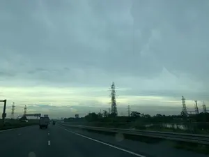 Bức ảnh được chụp từ một phương tiện đang di chuyển trên một tuyến đường cao tốc có nhiều làn xe. Bầu trời u ám, đầy mây, có thể là vào buổi sáng sớm hoặc chiều tà. Trên đường có các phương tiện khác đang lưu thông, bao gồm cả xe tải. Dọc theo đường là dải lan can bảo vệ. Bên kia lan can là một khung cảnh với cây cối, một con sông hoặc kênh nước, và rất nhiều cột điện cao thế bằng thép. Có một người dường như đang ngồi bên vệ đường, gần lan can và bờ sông. Ở phía xa có các biển báo giao thông nhưng không thể đọc rõ nội dung.。（日本語訳）。写真は、複数の車線がある高速道路を走行中の車両から撮影されたものです。空は曇っており、雲が多く、早朝か夕暮れ時かもしれません。道路にはトラックを含む他の車両が走行しています。道路に沿ってガードレールがあります。ガードレールの向こう側には、木々、川または水路、そして多くの鋼鉄製の高圧送電鉄塔がある風景が広がっています。路肩、ガードレールと川岸の近くに人が座っているようです。遠くには交通標識がありますが、内容ははっきりと読み取れません。