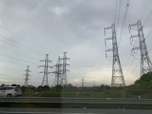 Bức ảnh được chụp từ bên trong một phương tiện đang di chuyển, nhìn ra cảnh quan bên đường. Nổi bật trong ảnh là một hệ thống gồm nhiều cột điện cao thế bằng kim loại và các đường dây điện chạy dọc theo một con đường. Bầu trời u ám và đầy mây. Ở phía trước là một dải lan can bảo vệ đường bộ. Bên kia lan can là một khu vực đất có cỏ và cây cối. Một chiếc ô tô màu trắng có thể được nhìn thấy ở phía bên trái. Không có văn bản hay biển báo nào có thể đọc được trong ảnh.。（日本語訳）。写真の中で際立っているのは、道路に沿って走る、多数の金属製高圧鉄塔と送電線からなる系統です。空はどんよりとしており、雲に覆われています。手前には道路のガードレールがあります。ガードレールの向こう側は、草や木が生えている土地です。左側には白い車が見えます。写真には、読み取れる文字や標識はありません。