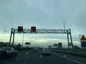 Bức ảnh chụp khung cảnh trên một con đường cao tốc nhiều làn xe, nhìn từ bên trong một chiếc ô tô đang di chuyển. Bầu trời nhiều mây, có thể là vào lúc sáng sớm hoặc chiều muộn. Nhiều xe ô tô đang lưu thông trên đường.。Có một giàn kết cấu thép lớn bắc ngang qua đường, trên đó có gắn các biển báo điện tử LED đang hiển thị các biểu tượng mũi tên màu đỏ.。Dọc theo lề đường bên phải và ở xa có các biển báo và biển quảng cáo với các nội dung sau:。- Biển quảng cáo: "RAPIDO GERMAN QUALITY", "KANSAI", "KY NGUYEN PASSION & PERFECTION".。- Biển báo khẩn cấp màu xanh lá cây: "KHẨN CẤP EMERGENCY", "S.O.S", và số điện thoại "1900599959".。- Biển báo giao thông màu xanh lam: "CHIỀU CAO AN TOÀN 7 m".。- Biển báo nguy hiểm hình tam giác màu vàng có biểu tượng tia sét, cảnh báo khu vực có điện cao thế.。Hai bên đường có cây cối, cột đèn đường và cột điện. Phía xa có thể thấy hình bóng của các tòa nhà.。（日本語訳）。複数の車線がある高速道路の風景を、走行中の車内から撮影した写真です。空は曇っており、早朝か夕方遅くかもしれません。多くの自動車が道路を走行しています。道路を横切る大きな鉄骨構造の架台があり、その上には赤い矢印の記号を表示しているLED電子標識が設置されています。右側の路肩沿いと遠方には、以下の内容の標識や広告看板があります。- 広告看板：「RAPIDO GERMAN QUALITY」、「KANSAI」、「KY NGUYEN PASSION & PERFECTION」。- 緑色の緊急標識：「KHẨN CẤP EMERGENCY」、「S.O.S」、および電話番号「1900599959」。- 青色の交通標識：「CHIỀU CAO AN TOÀN 7 m」。- 稲妻の記号がある黄色の三角形の危険標識は、高圧電線のある区域を警告しています。道路の両側には樹木、街灯、電柱があります。遠方には建物の影が見えます。