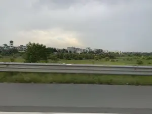 Bức ảnh được chụp từ một phương tiện đang di chuyển, cho thấy khung cảnh bên đường. Ở tiền cảnh là một phần của con đường nhựa và một dải lan can bằng kim loại. Phía sau lan can là một khu đất rộng với cỏ và cây cối xanh tươi. Ở phía xa là một khu dân cư với nhiều ngôi nhà san sát nhau, mang kiến trúc đặc trưng của vùng ngoại ô Việt Nam. Bầu trời u ám và đầy mây. Không có hoạt động cụ thể nào của con người hay bất kỳ văn bản nào có thể đọc được trong ảnh.。（日本語訳）。写真は移動中の乗り物から撮影されたもので、道端の風景が写っている。前景には、アスファルトの道路の一部と金属製のガードレールがある。ガードレールの後ろには、青々とした草木が生い茂る広大な土地が広がっている。遠くには、ベトナムの郊外特有の建築様式を持つ家々が密集した住宅街がある。空はどんよりとしており、雲に覆われている。写真には、特定の人物の活動や読み取れる文字は写っていない。