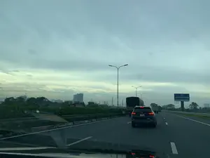 Trên một biển quảng cáo lớn màu xanh dương ở bên phải đường có dòng chữ 'XI MĂNG XUÂN THÀNH'. Khung cảnh được chụp từ trong một chiếc ô tô đang di chuyển trên đường cao tốc có nhiều làn xe. Có nhiều phương tiện khác đang tham gia giao thông, bao gồm một chiếc ô tô SUV màu đen ở ngay phía trước và một vài chiếc xe tải. Dọc theo con đường là hệ thống đèn chiếu sáng công cộng và cây xanh. Ở phía xa là hình ảnh của một khu đô thị với nhiều tòa nhà cao tầng và các công trình xây dựng có cần cẩu. Bầu trời u ám và nhiều mây.。（日本語訳）。道路の右側にある大きな青い広告看板に「XI MĂNG XUÂN THÀNH」という文字があります。この風景は、複数の車線がある高速道路を走行中の車内から撮影されたものです。他にも多くの車両が通行しており、すぐ前には黒いSUV車が一台、そして数台のトラックが含まれています。道路沿いには街灯や緑の木々があります。遠方には、多くの高層ビルやクレーンのある建設現場がある都市部の光景が見えます。空はどんよりとしており、雲が多いです。