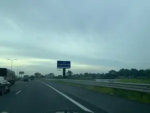 Bản ghi văn bản từ biển quảng cáo: Phía trên cùng là logo 'XUAN THANH GROUP'. Dòng chữ lớn nhất là 'XI MĂNG XUÂN THÀNH'. Dòng chữ nhỏ bên dưới là 'Dây chuyền FLSmidth Đan Mạch - Tiêu chuẩn xuất khẩu quốc tế'.。Mô tả cảnh quan và hoạt động: Hình ảnh được chụp từ bên trong một chiếc ô tô đang di chuyển trên đường cao tốc có nhiều làn xe. Nhiều phương tiện khác, bao gồm cả ô tô con và xe tải, cũng đang tham gia giao thông. Bầu trời có nhiều mây và thời tiết có vẻ âm u. Bên lề đường có một biển quảng cáo lớn cho Xi măng Xuân Thành. Phía sau dải phân cách kim loại là một khu vực có cây cối và một vùng nước như sông hoặc hồ. Xa xa là một số tòa nhà và nhà dân.。（日本語訳）。広告看板のテキスト記録：最上部は「XUAN THANH GROUP」のロゴ。最も大きな文字は「XI MĂNG XUÂN THÀNH」。その下の小さな文字は「デンマークFLSmidth社の生産ライン - 国際輸出基準」。風景と状況の説明：画像は、複数車線のある高速道路を走行中の車内から撮影されたものです。乗用車やトラックを含む他の多くの車両も通行しています。空は曇っており、天気はどんよりしているようです。道路脇にスアンタインセメントの大きな広告看板があります。金属製の中央分離帯の後ろには、木々や、川か湖のような水域があります。遠くにはいくつかのビルや民家が見えます。