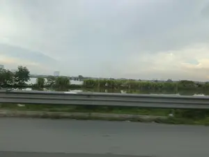 Bức ảnh ghi lại khung cảnh nhìn từ một phương tiện đang di chuyển trên đường. Ở tiền cảnh là mặt đường nhựa và một dãy lan can bảo vệ bằng kim loại. Ngay phía sau lan can là một khu vực mặt nước rộng lớn, có thể là hồ, ao hoặc một cánh đồng ngập nước, với cây cối và bụi rậm mọc ven bờ. Ở phía xa, đường chân trời hiện lên với bóng dáng của các tòa nhà cao tầng, cho thấy khu vực này nằm ở vùng ngoại ô hoặc gần một đô thị. Bầu trời u ám và có nhiều mây. Không có hoạt động nào của con người được quan sát, cũng không có bất kỳ văn bản hay biển báo nào trong ảnh.。（日本語訳）。写真は、道路を移動中の乗り物から見た風景を記録したものです。前景には、アスファルトの路面と一列の金属製ガードレールがあります。ガードレールのすぐ後ろには、湖、池、または冠水した畑である可能性のある広大な水域があり、岸辺には木々や茂みが生えています。遠くには、高層ビルのシルエットが地平線に現れており、この地域が郊外または都市の近くにあることを示しています。空はどんよりとしており、雲が多いです。人間の活動は観察されず、写真にはいかなる文字や標識もありません。