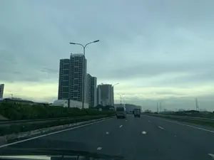 Bức ảnh được chụp từ bên trong một chiếc ô tô đang di chuyển trên một tuyến đường cao tốc nhiều làn xe. Khung cảnh là một khu đô thị hiện đại với nhiều tòa nhà chung cư cao tầng san sát. Trên nóc của tòa nhà cao nhất có một biển hiệu với chữ viết. Các phương tiện giao thông, bao gồm một xe khách nhỏ và một xe tải, đang lưu thông trên đường. Có một dải phân cách trồng cây xanh ở giữa. Dọc theo con đường là các cột đèn đường. Ở phía xa bên phải có thể thấy các cột điện cao thế. Bầu trời u ám và đầy mây.。（日本語訳）。写真は、複数車線の高速道路を走行中の車内から撮影されたものです。風景は、多くの高層マンションが密集する近代的な都市部です。最も高い建物の屋上には、文字が書かれた看板があります。小型バスやトラックを含む車両が道路を走行しています。中央には緑が植えられた中央分離帯があります。道路沿いには街灯が立っています。遠くの右側には、高圧電線の鉄塔が見えます。空はどんよりとしており、雲に覆われています。