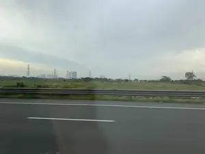 Đây là khung cảnh được chụp từ một phương tiện đang di chuyển trên đường cao tốc, nhìn ra ngoài cửa sổ. Ở tiền cảnh là mặt đường nhựa và một lan can bảo vệ. Phía sau lan can là một cánh đồng cỏ rộng lớn. Ở phía xa, có thể thấy nhiều cột điện cao thế và các tòa nhà chung cư cao tầng của một khu đô thị hiện ra mờ ảo. Bầu trời u ám và đầy mây. Không có hoạt động cụ thể nào của con người diễn ra, ngoại trừ việc di chuyển trên đường. Không có bất kỳ văn bản hay chữ viết nào trong ảnh.。（日本語訳）。これは、高速道路を走行中の乗り物から窓の外を撮影した風景です。手前にはアスファルトの路面とガードレールがあります。ガードレールの後ろには広大な草原が広がっています。遠くには、多くの高圧電線の鉄塔と、ぼんやりと現れる都市部の高層マンションが見えます。空はどんよりとしており、雲に覆われています。道路を移動していることを除いて、特定の人間活動は見られません。写真にはいかなるテキストや文字もありません。