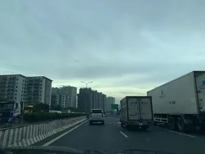 Bản ghi văn bản từ hình ảnh:。Trên biển báo giao thông lớn màu xanh lá cây ở phía xa có các dòng chữ: "PHÁP VÂN", "CẦU GIẼ", "EXPRESSWAY", "KẾT THÚC", "END", "500m".。Trên thùng của một chiếc xe tải nhỏ có logo "TST".。Trên thùng của một chiếc xe tải lớn màu trắng có dòng chữ "Hệ Thống Chuyên Cung Cấp..." và số điện thoại "Hotline" nhưng một phần bị che khuất.。Trên cửa một chiếc xe tải màu xanh đỗ bên lề đường có chữ "IN DẶM DINIKAN" nhưng không rõ nét.。Mô tả cảnh quan và hoạt động:。Đây là khung cảnh trên một tuyến đường cao tốc nhiều làn xe tại Việt Nam, có thể là đoạn gần cuối của đường cao tốc Pháp Vân - Cầu Giẽ như được chỉ dẫn trên biển báo. Hoạt động chính là các phương tiện giao thông đang di chuyển. Trong ảnh có nhiều loại xe, bao gồm một chiếc ô tô con màu bạc, một xe tải thùng nhỏ và một xe tải thùng lớn màu trắng đang chạy trên đường. Con đường được phân cách bởi dải phân cách bê tông sơn sọc trắng đỏ. Phía xa là cảnh quan đô thị với nhiều tòa nhà chung cư cao tầng, một số tòa nhà có vẻ đang trong quá trình xây dựng. Bầu trời u ám và nhiều mây.。（日本語訳）。画像からのテキスト記録：。遠くにある大きな緑色の交通標識には、「PHÁP VÂN」、「CẦU GIẼ」、「EXPRESSWAY」、「KẾT THÚC」、「END」、「500m」の文字があります。小型トラックの荷台には「TST」のロゴがあります。大きな白いトラックの荷台には、「Hệ Thống Chuyên Cung Cấp...」という文字と「Hotline」の電話番号がありますが、一部が隠されています。路肩に停まっている青いトラックのドアには「IN DẶM DINIKAN」という文字がありますが、不鮮明です。風景と活動の描写：。これはベトナムの複数車線ある高速道路の光景で、標識に示されているように、ファップバン-カウゼー高速道路の終点付近の区間である可能性があります。主な動きは、車両が走行していることです。道路は、赤と白の縞模様に塗装されたコンクリートの中央分離帯で仕切られています。遠方には、多くの高層マンションが建ち並ぶ都市の景観が広がっており、一部の建物は建設中のようです。空はどんよりと曇っています。