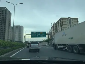 Bức ảnh chụp cảnh giao thông trên một đường cao tốc, nhìn từ trong xe ô tô. Trên đường có nhiều phương tiện đang lưu thông, bao gồm một chiếc xe SUV màu bạc ở phía trước và một xe tải container màu trắng ở làn bên phải. Ở phía trên có một biển báo chỉ dẫn giao thông lớn màu xanh lá cây. Biển báo chỉ hướng đi thẳng theo 'QL.1A PHÁP VÂN QUỐC LỘ 1A' và hướng rẽ vào 'CT.20 TP. HÀ NỘI ĐẠI LỘ THĂNG LONG'. Hai bên đường là các tòa nhà cao tầng; đáng chú ý là một tòa nhà lớn bên trái đang trong quá trình xây dựng và một tòa chung cư đã có người ở bên phải. Trên thành của xe tải container có in logo và dòng chữ quảng cáo: 'Hệ thống Chuyên Cung Cấp MỰA TƯƠI, MỰA KHÔ Toàn Quốc' cùng với số điện thoại 'Hotline: 0944.575.555 - 0915.088.828'. Khung cảnh cho thấy một tuyến đường huyết mạch ở khu vực đô thị, với bầu trời có nhiều mây.。（日本語訳）。自動車の中から撮影された、ある高速道路上の交通風景の写真です。道路上には多くの車両が通行しており、前方には一台の銀色のSUV、右車線には一台の白色のコンテナトラックが含まれています。上方には、大きな緑色の交通案内標識があります。標識は、直進方向が「QL.1A PHÁP VÂN QUỐC LỘ 1A」、分岐方向が「CT.20 TP. HÀ NỘI ĐẠI LỘ THĂNG LONG」であることを示しています。道路の両側には高層ビルがあります。特筆すべきは、左側の大きなビルが建設中であり、右側には入居済みの集合住宅があることです。コンテナトラックの側面には、「全国 生MỰA、干しMỰA 専門供給システム」というロゴと広告文が、「Hotline: 0944.575.555 - 0915.088.828」という電話番号と共に印刷されています。この光景は、都市部の幹線道路を示しており、空には多くの雲が浮かんでいます。