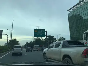 Bức ảnh được chụp từ bên trong một chiếc ô tô, ghi lại cảnh giao thông trên một tuyến đường cao tốc ở Việt Nam. Ở trung tâm bức ảnh là một biển báo lớn màu xanh lá cây, trên đó có ghi các thông tin sau:。'CT.01 PHÁP VÂN - CẦU GIẼ'。'EXPRESSWAY'。'KẾT THÚC'。'END'。Biển báo này cho biết đây là điểm kết thúc của đường cao tốc Pháp Vân - Cầu Giẽ. Trên đường có nhiều phương tiện đang lưu thông, bao gồm một chiếc xe SUV màu bạc ở phía trước, một chiếc xe bán tải màu trắng ở bên phải, và các xe ô tô khác. Bên phải đường là một tòa nhà cao tầng hiện đại với mặt tiền bằng kính. Bối cảnh phía sau là cây cối, cột điện cao thế và bầu trời u ám, nhiều mây.。（日本語訳）。写真は自動車の中から撮影されたもので、ベトナムのある高速道路の交通風景を記録しています。写真の中央には大きな緑色の看板があり、そこには以下の情報が書かれています。「CT.01 ファップヴァン - カウゼー」。「EXPRESSWAY」。「KẾT THÚC（終了）」。「END」。この看板は、ここがファップヴァン-カウゼー高速道路の終点であることを示しています。道路上には多くの車両が通行しており、前方には銀色のSUV、右側には白色のピックアップトラック、そしてその他の自動車が含まれています。道路の右側には、ガラス張りのファサードを持つ近代的な高層ビルがあります。