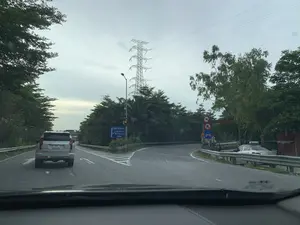 Bức ảnh ghi lại khung cảnh giao thông trên một con đường có ngã rẽ, được chụp từ góc nhìn bên trong một chiếc ô tô. Bầu trời u ám. Phía trước là một chiếc xe SUV màu bạc đang di chuyển trên làn đường bên trái. Tại ngã rẽ, có một biển báo chỉ dẫn lớn màu xanh lam chỉ hướng đi về bên trái, ghi các điểm đến bao gồm 'TRUNG TÂM HÀ NỘI', 'CẦU THANH TRÌ', 'BẮC THĂNG LONG', 'NỘI BÀI'. Ở lối rẽ bên phải, có các biển báo cấm xe mô tô và người đi bộ. Cảnh vật xung quanh bao gồm nhiều cây xanh rậm rạp hai bên đường và một cột điện cao thế lớn. Phía xa bên phải có một chiếc taxi màu trắng đang đỗ, một chiếc xe tải và một người đi xe máy.。（日本語訳）。写真は、分岐のある道路の交通風景を、一台の車内からの視点で撮影したものである。空は曇っている。前方には、左車線を走行している銀色のSUVが一台いる。分岐点には、左方向への行き先を示す大きな青い案内標識があり、「ハノイ中心部」「タインチ橋」「バクタンロン」「ノイバイ」を含む目的地が記載されている。右の分岐路には、バイクと歩行者を禁止する標識がある。周囲の景色には、道路の両脇に生い茂る多くの緑の木々と、一本の大きな高圧電線の鉄塔が含まれている。右手遠方には、一台の白いタクシーが停車しており、一台のトラックとバイクに乗った一人がいる。