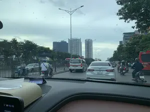 Bức ảnh được chụp từ bên trong một chiếc ô tô, ghi lại cảnh giao thông trên một con đường đô thị tại Việt Nam. Thời tiết có vẻ u ám và có những giọt nước trên kính chắn gió, cho thấy trời đang hoặc vừa mới mưa.。Hoạt động chính là các phương tiện đang tham gia giao thông. Trên đường có nhiều ô tô và xe máy. Phía trước có một chiếc xe cứu thương màu trắng với dòng chữ 'CẤP CỨU 24/24' và 'HOTLINE: 0918 366 115'. Bên cạnh đó là các xe ô tô con và nhiều người đi xe máy, hầu hết đều đội mũ bảo hiểm. Một chiếc xe khách màu đỏ ở phía bên phải có chữ 'HÀNH KHÁCH' và một phần tên nhà xe 'SaoViệt'.。Ở hậu cảnh là các tòa nhà cao tầng. Một tòa nhà có logo 'MB'. Một tòa nhà khác có chữ 'SUPERMARKET' không rõ ràng. Một công trình xây dựng cao tầng đang thi công với cần cẩu ở trên đỉnh. Dọc hai bên đường là cây xanh, hàng rào và các cột đèn đường.。Ở phía trước, có thể thấy một phần bảng điều khiển của ô tô và một hộp đựng khăn giấy. Trên hộp khăn giấy có một vật trang trí nhỏ ghi chữ 'Khải'.。（日本語訳）。写真は車内から撮影されたもので、ベトナムのある都市の道路の交通風景を記録しています。天候は曇っているようで、フロントガラスには水滴が付いており、雨が降っているか、降ったばかりであることを示しています。主な活動は、交通に参加している車両です。道路には多くの自動車やバイクがいます。前方には、「CẤP CỨU 24/24」（24時間救急）と「HOTLINE: 0918 366 115」という文字が書かれた白い救急車が一台あります。その隣には乗用車や多くのバイクに乗る人々がおり、ほとんどがヘルメットを着用しています。背景には高層ビルがあります。あるビルには「MB」のロゴがあります。別のビルには、はっきりとしない「SUPERMARKET」の文字があります。頂上にクレーンがある高層建築物が建設中です。道路の両側には緑の木々、フェンス、街灯があります。前方には、車のダッシュボードの一部とティッシュボックスが見えます。ティッシュボックスの上には、「Khải」と書かれた小さな装飾品があります。