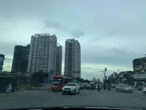 Bức ảnh chụp cảnh quan đô thị tại một ngã tư đông đúc, được chụp từ bên trong một chiếc ô tô qua kính chắn gió có dính vài giọt nước. Bầu trời u ám.。【Chữ viết được ghi lại】。*   Trên nóc tòa nhà cao tầng bên trái: "IMPERIAL", "M SUPERMARKET".。*   Trên nóc tòa nhà cao tầng bên phải: Logo "MB".。*   Trên xe khách màu đỏ: "VIP", "TÂN", "2023", "NAM ĐỊNH - CỔ LỄ HẢI NINH", "ĐOÀN KẾT".。*   Trên biển hiệu bên phải: "NAM SƠN E...", "NAM SƠN".。【Mô tả cảnh vật và hoạt động】。*   **Địa điểm:** Một ngã tư đường phố lớn ở khu vực thành thị, có nhiều tòa nhà cao tầng và mật độ giao thông cao. Bên trái có một công trình xây dựng đang thi công, được rào chắn và che lưới đen.。*   **Hoạt động:** Các phương tiện bao gồm ô tô, xe máy và xe khách đang tham gia giao thông trên đường. Một số phương tiện đang dừng chờ đèn tín hiệu. Một người phụ nữ mặc váy xanh đang đi bộ ở lề đường bên trái.。*   **Đối tượng:** Hai tòa nhà chung cư/văn phòng cao tầng là điểm nhấn chính của khung cảnh. Dưới đường là dòng xe cộ tấp nập, nổi bật là một chiếc xe khách màu đỏ và một chiếc ô tô SUV màu trắng. Nhiều người đang điều khiển xe máy. Cơ sở hạ tầng đường phố như đèn giao thông và cột đèn cũng hiện diện rõ.。（日本語訳）。混雑した交差点の都市景観を、水滴がいくつか付着した車のフロントガラス越しに車内から撮影した写真。空は曇っている。【記録された文字】。*   左側の高層ビルの屋上：「IMPERIAL」、「M SUPERMARKET」。*   右側の高層ビルの屋上：「MB」のロゴ。*   赤いバス：「VIP」、「TÂN」、「2023」、「NAM ĐỊNH - CỔ LỄ HẢI NINH」、「ĐOÀN KẾT」。*   右側の看板：「NAM SƠN E...」、「NAM SƠN」。【風景と活動の説明】。*   **場所：**都市部にある大きな交差点で、多くの高層ビルがあり交通量が多い。左側には、フェンスで囲まれ黒いネットで覆われた建設中の工事現場がある。*   **活動：**自動車、バイク、バスなどの車両が道路を通行している。一部の車両は信号待ちで停止している。青いスカートを履いた女性が左側の路肩を歩いている。*   **対象物：**2つの高層マンション/オフィスビルが風景の主要な見どころとなっている。道路にはひっきりなしに車が流れており、赤いバスと白いSUV車が目立っている。多くの人がバイクを運転している。信号機や街灯などの道路インフラもはっきりと存在している。