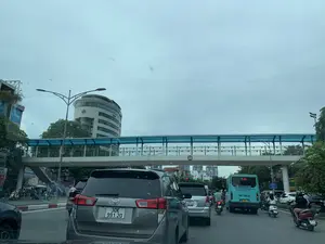 Quang cảnh giao thông trên một con đường đô thị sầm uất vào một ngày trời nhiều mây. Dòng xe cộ đông đúc gồm nhiều ô tô và xe máy đang di chuyển trên đường. Nổi bật ở phía trước là một chiếc ô tô Toyota Innova màu xám. Bên phải là một chiếc xe buýt màu xanh lam và nhiều người đi xe máy. Phía trên đường là một cây cầu vượt dành cho người đi bộ có kết cấu bằng kim loại và mái che màu xanh. Trong nền là một tòa nhà văn phòng cao tầng với kiến trúc hiện đại.。Các thông tin bằng chữ viết có thể nhận dạng bao gồm:。- Trên xe buýt: "CÔNG TY CP XE KHÁCH HÀ NỘI", "HOTLINE: 0243 873 2663", và số tuyến "146" trên bảng điện tử.。- Biển báo trên cầu vượt: Biển báo giới hạn chiều cao "4.75m".。- Biển báo bên đường: Một biển báo màu xanh ghi chữ "ĐÈN ĐỎ".。- Biển hiệu cửa hàng: Một phần biển hiệu ghi "AO MAI AUDIO".。- Chữ trên xe ô tô: "INNOVA", "TOYOTA MY DINH".。（日本語訳）。曇りの日の、賑やかな都市の道路における交通風景。多くの自動車やバイクからなる混雑した車両の流れが道路上を移動している。手前に目立っているのは、灰色のトヨタ・イノーバだ。右側には青いバスと多くのバイクに乗った人々がいる。道路の上には、金属構造で青い屋根の歩行者用陸橋がある。背景には、近代的な建築の高層オフィスビルがある。識別可能な文字情報には以下が含まれる：。- バスの表示：「CÔNG TY CP XE KHÁCH HÀ NỘI」（ハノイ旅客自動車株式会社）、「HOTLINE: 0243 873 2663」、および電光掲示板の路線番号「146」。- 陸橋の標識：高さ制限標識「4.75m」。- 道路脇の標識：「ĐÈN ĐỎ」（赤信号）と書かれた青い標識。- 店舗の看板：「AO MAI AUDIO」と書かれた看板の一部。- 自動車の文字：「INNOVA」、「TOYOTA MY DINH」。