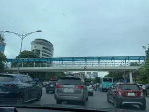 Bối cảnh là một con đường đô thị đông đúc với nhiều phương tiện giao thông như ô tô, xe máy và xe buýt đang di chuyển chậm hoặc dừng lại, có thể là do ùn tắc. Phía trên đường có một cây cầu vượt dành cho người đi bộ với kết cấu thép và mái che màu xanh lam. Các phương tiện bao gồm nhiều loại ô tô khác nhau và một chiếc xe buýt màu xanh lam. Ở phía sau là các tòa nhà cao tầng, nổi bật là một tòa nhà văn phòng có kiến trúc hình trụ hiện đại. Các thông tin bằng chữ có thể đọc được bao gồm: trên cầu vượt có biển báo giới hạn chiều cao '4,75m'; trên chiếc xe buýt màu xanh lam có số tuyến '146', dòng chữ 'CÔNG TY CP XE KHÁCH HÀ NỘI', và 'HOTLINE: 0243 873 2663'; trên một chiếc ô tô màu xanh dương có dán sticker 'Ô DÂN CHỦ'; một biển báo bên đường có chữ 'ĐẾN ĐÓ CHÚ Ý'; và một biển hiệu khác có số '688'.。（日本語訳）。背景は、自動車、バイク、バスなどの多くの交通車両が、おそらく渋滞が原因で、ゆっくりと移動または停止している、混雑した都市の道路です。道路の上には、鉄骨構造で青い屋根の歩行者用陸橋があります。車両には、様々な種類の自動車と一台の青いバスが含まれています。後方には高層ビルがあり、特に目立つのは近代的な円筒形の建築様式を持つオフィスビルです。読み取れる文字情報には、以下が含まれます：陸橋には「4,75m」の高さ制限標識があります。青いバスには、路線番号「146」、「CÔNG TY CP XE KHÁCH HÀ NỘI」の文字、そして「HOTLINE: 0243 873 2663」があります。青い自動車には「Ô DÂN CHỦ」のステッカーが貼られています。道路脇の標識には「ĐẾN ĐÓ CHÚ Ý」の文字があります。そして、別の看板には「688」という数字があります。