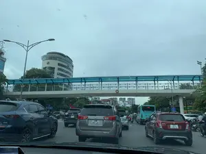 Bối cảnh là một con đường đông đúc trong một thành phố, được chụp từ bên trong một chiếc ô tô. Có rất nhiều phương tiện đang dừng lại hoặc di chuyển chậm, cho thấy tình trạng tắc nghẽn giao thông. Các phương tiện bao gồm nhiều loại ô tô và xe máy. Một chiếc xe buýt màu xanh lá cây đang ở làn đường bên phải. Phía trên đường là một cây cầu vượt dành cho người đi bộ có mái che màu xanh lam. Ở phía xa là các tòa nhà cao tầng, trong đó có một tòa nhà văn phòng hiện đại với kiến trúc hình tròn. Hoạt động chính đang diễn ra là các phương tiện đang tham gia giao thông trên đường phố vào một ngày trời nhiều mây.。Các dòng chữ có thể đọc được trong ảnh:。- Trên cầu vượt có biển báo giới hạn chiều cao hình tròn ghi "4,75m".。- Trên xe buýt màu xanh có số tuyến "146", và dòng chữ "CÔNG TY CP XE KHÁCH HÀ NỘI", "HOTLINE: 0243 873 2663".。- Một biển báo giao thông màu xanh có chữ "ĐIỂM ĐỖ" và "CHÚ Ý".。- Một biển hiệu trên tòa nhà có chữ "GB8" và số điện thoại "1800...".。- Một nhãn dán trên xe ô tô phía bên trái có chữ "Ô DẪN CHÚ", tuy nhiên chữ này không rõ ràng.。（日本語訳）。背景は、ある都市の混雑した道路で、車内から撮影されたものです。多くの車両が停止または徐行しており、交通渋滞の状況を示しています。車両には、多くの種類の自動車やバイクが含まれています。右側の車線には緑色のバスがいます。道路の上には、青い屋根の歩道橋があります。遠くには高層ビルがあり、その中には円形の建築様式を持つ近代的なオフィスビルがあります。主な活動は、曇りの日に車両が道路交通に参加していることです。写真で読み取れる文字：。- 歩道橋には、「4,75m」と書かれた円形の高さ制限標識があります。- 緑色のバスには、路線番号「146」と、「CÔNG TY CP XE KHÁCH HÀ NỘI」（ハノイ旅客自動車株式会社）、「HOTLINE: 0243 873 2663」という文字があります。- 青い交通標識に「ĐIỂM ĐỖ」（駐車場）と「CHÚ Ý」（注意）という文字があります。- 建物にある看板に「GB8」という文字と電話番号「1800...」があります。- 左側の車にあるステッカーに「Ô DẪN CHÚ」という文字がありますが、この文字は不鮮明です。