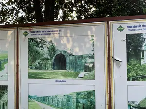 Bức ảnh chụp một bảng thông tin lớn đặt ngoài trời, dưới một tán cây. Trên bảng có các dòng chữ:。- Tiêu đề chính: "TRUNG TÂM BẢO TỒN DI SẢN THÀNH NHÀ HỒ" và bên dưới là "CONSERVATION CENTER FOR HO CITADEL".。- Chú thích cho bức ảnh trung tâm: "CỔNG ĐÔNG".。- Trên một ô thông tin khác bên phải: "TRUNG TÂM BẢO TỒN" và "CONSERVATION CEN".。Bối cảnh là tại khu di tích Thành Nhà Hồ. Bảng thông tin này có nhiệm vụ giới thiệu các hình ảnh về di tích cho du khách. Các vật thể chính bao gồm:。- Bảng thông tin: Có khung màu nâu, được chia thành nhiều ô, mỗi ô là một bức ảnh về Thành Nhà Hồ.。- Các bức ảnh: Bức ảnh nổi bật nhất ở giữa chụp Cổng Đông, một cổng thành cổ được xây bằng những khối đá lớn với kiến trúc mái vòm. Các bức ảnh khác cho thấy các đoạn tường thành bằng đá.。- Cây cối: Phía trên bảng hiệu là một cây lớn có nhiều lá, đổ bóng xuống bảng.。（日本語訳）。写真は、木陰の屋外に設置された大きな案内板を写しています。案内板には以下の文字が書かれています：。- メインタイトル：「TRUNG TÂM BẢO TỒN DI SẢN THÀNH NHÀ HỒ」、その下に「CONSERVATION CENTER FOR HO CITADEL」。- 中央の写真のキャプション：「CỔNG ĐÔNG」。- 右側の別の情報欄には：「TRUNG TÂM BẢO TỒN」と「CONSERVATION CEN」。背景は胡朝の城跡です。この案内板は、観光客に遺跡の写真を紹介する役割を担っています。主な被写体は以下の通りです：。- 案内板：茶色のフレームがあり、複数の区画に分かれており、各区画は胡朝の城の写真です。- 写真：中央の最も目立つ写真は東門を写したもので、大きな石のブロックで建てられたアーチ構造の古い城門です。他の写真は石の城壁の一部を示しています。- 樹木：看板の上には葉の多い大きな木があり、看板に影を落としています。