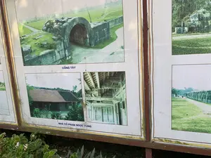 Bức ảnh chụp một bảng thông tin đã cũ, có khung kim loại, được đặt ngoài trời tại một khu di tích. Trên bảng có dán nhiều hình ảnh giới thiệu về các địa điểm trong khu di tích. Có thể đọc được các dòng chữ chú thích cho các bức ảnh: 'CỔNG TÂY' và 'NHÀ CỔ PHẠM NGỌC TÙNG'. Bức ảnh có chú thích 'CỔNG TÂY' mô tả một cổng thành cổ được xây bằng những khối đá lớn với kiến trúc hình vòm, xung quanh là bãi cỏ. Các bức ảnh có chú thích 'NHÀ CỔ PHẠM NGỌC TÙNG' cho thấy một ngôi nhà cổ truyền thống với mái ngói và một bức ảnh khác chụp chi tiết kết cấu gỗ với các họa tiết chạm khắc tinh xảo bên trong. Các hình ảnh khác trên bảng cho thấy các đoạn tường thành bằng đá và con đường mòn. Phía dưới bảng thông tin có một vài luống cây hoa nhỏ.。（日本語訳）。写真は、ある遺跡地区の屋外に設置された、金属フレームの古い案内板を写している。板の上には、遺跡地区内の各場所を紹介する多くの写真が貼られている。写真のキャプションの文字を読むことができる：「西門」と「ファム・ゴック・トゥン古民家」。「西門」というキャプションのある写真は、大きな石のブロックで建てられたアーチ構造の古い城門を描写しており、周りは芝生である。「ファム・ゴック・トゥン古民家」というキャプションのある写真群は、瓦屋根の伝統的な古民家と、内部の精巧な彫刻模様が施された木造構造の詳細を写した別の写真を示している。板上の他の写真は、石造りの城壁の一部と小道を示している。案内板の下側には、いくつかの小さな花壇がある。