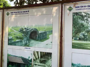 Bức ảnh chụp một bảng thông tin lớn đặt ngoài trời tại khu di tích Thành nhà Hồ. Trên bảng có tiêu đề 'TRUNG TÂM BẢO TỒN DI SẢN THÀNH NHÀ HỒ' và bản dịch tiếng Anh 'CONSERVATION CENTER FOR HO CITADEL', cùng với logo của di tích. Bảng thông tin trưng bày nhiều hình ảnh về các hạng mục của Thành nhà Hồ. Bức ảnh nổi bật nhất, được chú thích là 'CỔNG TÂY', cho thấy một cổng thành bằng đá đồ sộ với kiến trúc mái vòm, được xây từ những khối đá lớn, nằm giữa một bãi cỏ rộng. Các bức ảnh nhỏ hơn ở phía dưới và bên cạnh mô tả các chi tiết kiến trúc khác, bao gồm một ngôi nhà mái ngói truyền thống và kết cấu gỗ bên trong một công trình.。（日本語訳）。写真は、胡朝の城跡に屋外設置された大きな案内板を写しています。案内板には「TRUNG TÂM BẢO TỒN DI SẢN THÀNH NHÀ HỒ」というタイトルとその英訳「CONSERVATION CENTER FOR HO CITADEL」、そして遺跡のロゴがあります。案内板には、胡朝の城の各項目に関する多くの写真が展示されています。この城門は大きな石のブロックで造られ、広い芝生の中にあります。下部と側面にある小さな写真には、伝統的な瓦屋根の家や建造物内部の木造構造など、他の建築の詳細が描かれています。