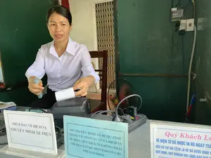 Bối cảnh là một quầy bán vé, có thể là tại một khu du lịch. Một nhân viên nữ mặc áo sơ mi trắng đang ngồi sau quầy, thực hiện giao dịch bán vé. Cô đang cầm tiền giấy trong một tay và tay kia đang lấy một chiếc vé được in ra từ máy in. Trên bàn làm việc có một máy in vé, một chiếc quạt điện màu đen, một ổ cắm điện hiệu SINO và nhiều bảng thông báo cho khách hàng.。Nội dung các bảng thông báo có thể đọc được như sau:。1. Bảng màu trắng: "ĐIỂM BÁN VÉ DỊCH VỤ THUYẾT MINH XE ĐIỆN".。2. Bảng màu xanh lam: "PHÍ THUYẾT MINH ĐÃ ĐƯỢC TÍNH TRONG VÉ THAM QUAN VÀ DỊCH VỤ XE ĐIỆN, QUÝ KHÁCH KHÔNG PHẢI CHI TRẢ BẤT KỂ MỘT KHOẢN CHI PHÍ NÀO KHÁC".。3. Bảng có chữ màu hồng (bị che một phần): "Quý Khách Lưu ý. VÉ ĐIỆN TỬ ĐÃ ĐƯỢC IN RÕ NGÀY THÁ... NẾU CÓ SỰ SAI LỆCH LÀ VÉ ĐÃ ĐƯỢC QUAY V... QUY ĐỊNH, QUÝ KHÁCH VUI LÒNG BÁO QUA...".。Phía sau người phụ nữ là một bức tường màu xanh lá cây và một cánh cửa xếp bằng sắt.。（日本語訳）。背景はチケット売り場で、おそらく観光地にあるものです。白いシャツを着た女性スタッフがカウンターの後ろに座り、チケットの販売取引を行っています。彼女は片方の手で紙幣を持ち、もう片方の手でプリンターから印刷されたチケットを取っています。仕事用の机の上には、チケットプリンター1台、黒い扇風機1台、SINOブランドのコンセント1つ、そして顧客向けの案内板が多数あります。読み取れる案内板の内容は以下の通りです。1. 白い板：「電気自動車 解説サービス チケット販売所」。2. 青い板：「解説料は観光チケットおよび電気自動車サービスに含まれておりますので、お客様はその他のいかなる費用もお支払いいただく必要はございません」。3. ピンク色の文字の板（一部が隠れている）：「お客様へのお知らせ。電子チケットには日付がはっきりと印刷されて... もし相違がある場合、そのチケットは...回転...規定...です。お客様は...までお知らせください...」。女性の後ろには緑色の壁と鉄製の折りたたみドアがあります。