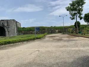 Nội dung văn bản trên các biển báo:。Biển chỉ dẫn cột cao có ba biển nhỏ màu xanh:。1. TT Bảo tồn Di sản Thành Nhà Hồ。2. Nhà vệ sinh / Toilets。3. Nhà trưng bày / Exhibitions (100m)。Biển chỉ dẫn khác ở phía xa hơn:。- BÃI ĐỖ XE。Mô tả cảnh quan và hoạt động:。Bức ảnh chụp một khung cảnh ngoài trời tại một khu di tích lịch sử, cụ thể là Thành nhà Hồ ở Thanh Hóa. Bên trái là một phần của một công trình kiến trúc cổ bằng đá lớn, là một cổng thành hoặc một đoạn tường thành. Một con đường trải sỏi dẫn vào bên trong khu vực.。Ở giữa bức ảnh có một cột biển báo chỉ đường cho du khách đến các địa điểm khác nhau: Trung tâm Bảo tồn Di sản Thành Nhà Hồ, nhà vệ sinh, và nhà trưng bày. Xa hơn một chút, có một biển báo khác chỉ lối vào bãi đỗ xe.。Xung quanh là cây xanh, bãi cỏ và một bờ tường thành được đắp bằng đất và gia cố bằng đá. Có một cột đèn đường trong khu vực. Bầu trời có mây. Không có người hay hoạt động nào đang diễn ra trong ảnh, cho thấy một không gian yên tĩnh tại khu di tích.。（日本語訳）。看板の表示内容：。高いポールにある案内標識には、3つの青い小さな看板があります。1. 胡朝の城跡遺産保存センター。2. トイレ / Toilets。3. 展示室 / Exhibitions (100m)。さらに遠くにある別の案内標識：。- 駐車場。風景と活動の説明：。写真は、歴史的遺跡、具体的にはタインホア省にある胡朝の城の屋外の風景を撮影したものです。左側には、大きな石でできた古代建築物の一部があり、それは城門または城壁の一部です。砂利道がエリアの内側へと続いています。写真の中央には、胡朝の城跡遺産保存センター、トイレ、展示室など、さまざまな場所へ観光客を案内する標識ポールがあります。少し離れたところには、駐車場への入り口を示す別の看板があります。周囲には緑の木々、芝生、そして土で盛られ石で補強された城壁があります。エリア内には街灯が一本あります。空は曇っています。写真には人や活動の様子はなく、遺跡の静かな空間が示されています。