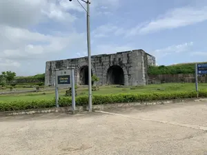 Bản ghi văn bản từ các biển báo trong hình:。1. Biển báo chỉ dẫn gần nhất (màu xanh lá): 'CỔNG NAM', 'South Gate', 'TRUNG TÂM BẢO TỒN DI SẢN THÀNH NHÀ HỒ', 'Conservation Center for Ho Citadel'.。2. Biển báo chỉ dẫn ở xa hơn (màu xanh dương): 'TT Bảo tồn Di sản Thành Nhà Hồ', 'Nhà vệ sinh / Toilets 100 m', 'Nhà trưng bày / Exhibitions 100 m', 'P' (Bãi đỗ xe).。Mô tả cảnh quan và hoạt động:。Bức ảnh chụp Cổng Nam của di tích lịch sử Thành nhà Hồ. Đây là một công trình kiến trúc bằng đá đồ sộ, cổ kính với ba cửa ra vào hình vòm lớn. Cổng được xây dựng từ những phiến đá lớn, xếp chồng lên nhau. Xung quanh cổng là bãi cỏ xanh và một số cây cảnh được trồng để trang trí. Phía trước là một khoảng sân hoặc đường đi đã được bê tông hóa. Bầu trời trong xanh và có mây. Không có hoạt động nào của con người được ghi lại, cho thấy một khung cảnh yên bình tại một địa điểm du lịch lịch sử.。（日本語訳）。画像内の看板のテキスト記録：。1. 最も近い案内看板（緑色）：「CỔNG NAM」、「South Gate」、「TRUNG TÂM BẢO TỒN DI SẢN THÀNH NHÀ HỒ」、「Conservation Center for Ho Citadel」。2. より遠くの案内看板（青色）：「TT Bảo tồn Di sản Thành Nhà Hồ」、「Nhà vệ sinh / Toilets 100 m」、「Nhà trưng bày / Exhibitions 100 m」、「P」（駐車場）。景観と活動の描写：。写真は歴史遺跡である胡朝の城壁の南門を撮影したものです。これは、3つの大きなアーチ型の出入り口を持つ、巨大で古風な石造りの建築物です。門は大きな石板を積み重ねて建設されています。門の周りは緑の芝生で、装飾のためにいくつかの観賞用の木が植えられています。正面にはコンクリートで舗装された広場または通路があります。空は青く澄んでおり、雲があります。人間の活動は記録されておらず、歴史的な観光地での静かな風景を示しています。